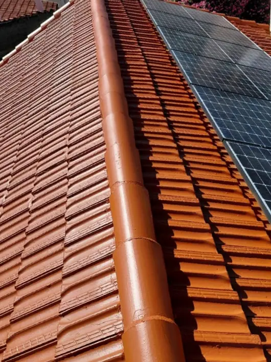 La même toiture nettoyée puis peinte en teinte terre cuite, faîtage repeint, mêmes panneaux photovoltaïques — HH Couverture